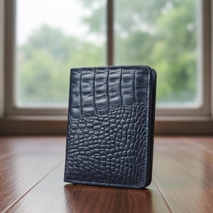 May include: A navy blue wallet with a crocodile skin texture. The wallet is rectangular and appears to be made of leather. The wallet is standing upright on a wooden surface.