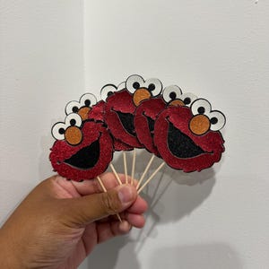 May include: A fan of red glitter Elmo face cutouts with black mouths, orange noses, and white and black eyes, attached to wooden sticks. The cutouts are held in a hand against a white background.