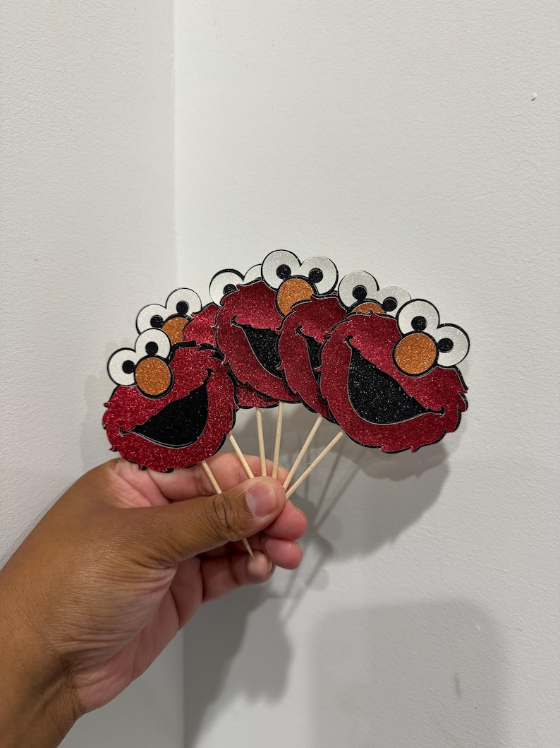 May include: A fan of red glitter Elmo face cutouts with black mouths, orange noses, and white and black eyes, attached to wooden sticks. The cutouts are held in a hand against a white background.