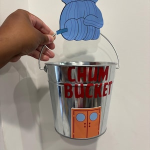 May include: A shiny metal bucket with the words "CHUM BUCKET" in red lettering. A blue cartoon hand holds the bucket's handle. The bucket has an orange door graphic with two blue circles.
