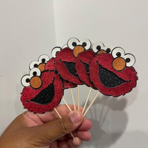May include: A hand holds several red glitter cupcake toppers shaped like a cartoon character with large white eyes, orange nose, and a black smiling mouth. The toppers are attached to wooden sticks.