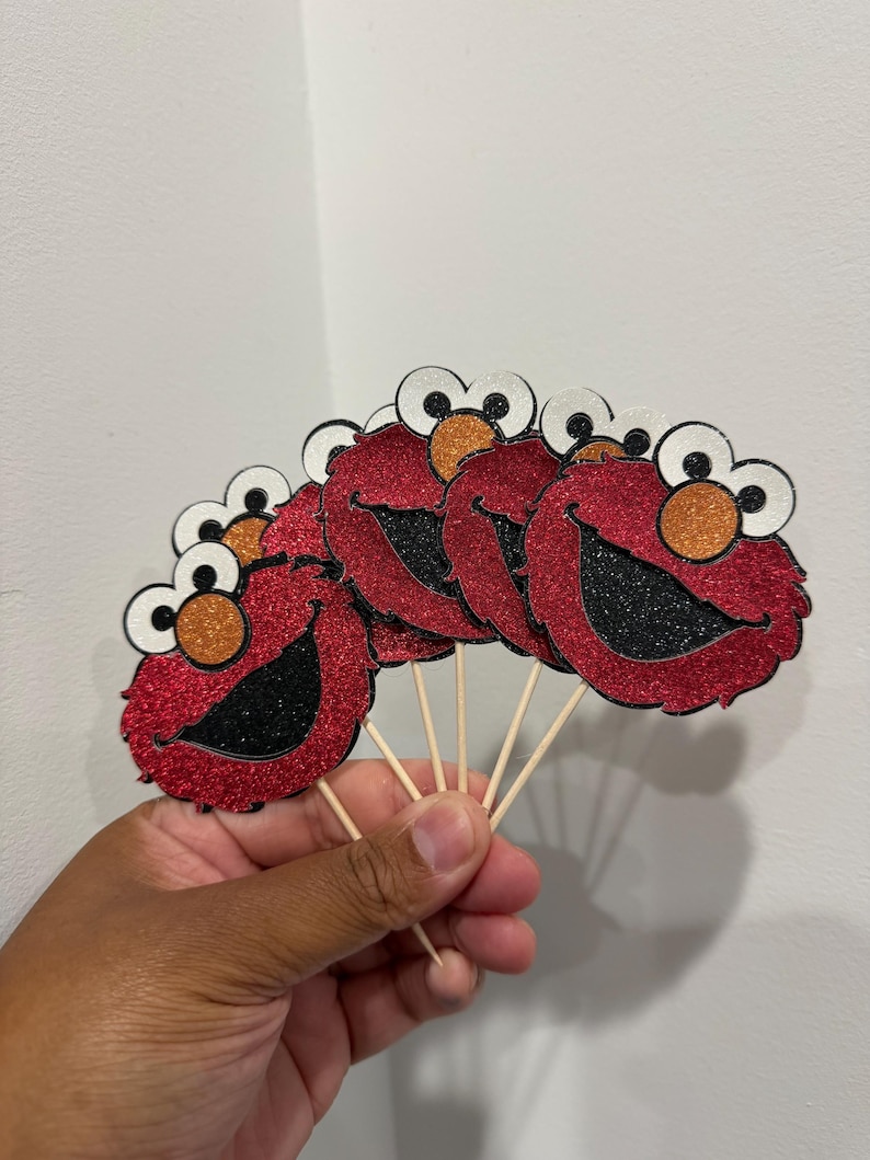 May include: A hand holds several red glitter cupcake toppers shaped like a cartoon character with large white eyes, orange nose, and a black smiling mouth. The toppers are attached to wooden sticks.