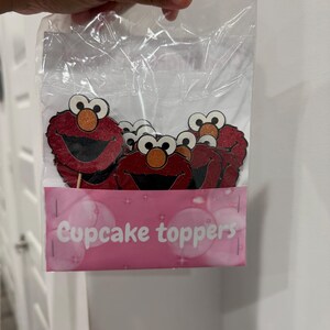 May include: A clear plastic bag containing several red Elmo cupcake toppers. The toppers have a glittery texture and are attached to wooden sticks. A pink label at the bottom reads "Cupcake toppers" in white text.