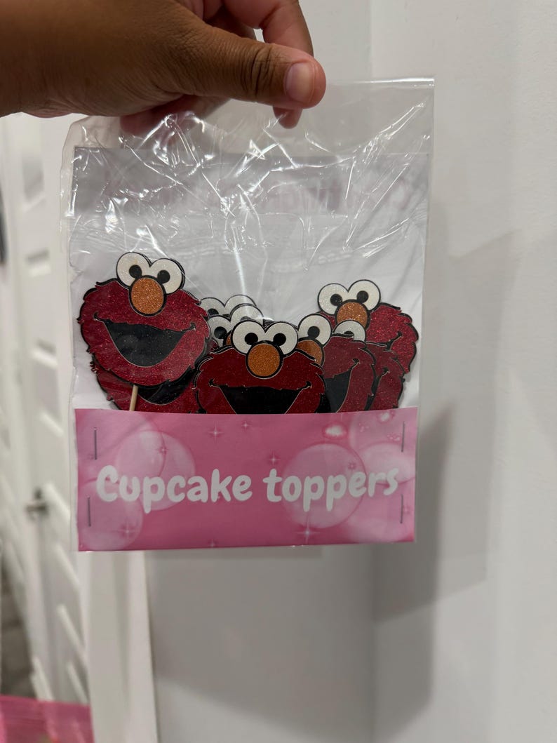 May include: A clear plastic bag containing several red Elmo cupcake toppers. The toppers have a glittery texture and are attached to wooden sticks. A pink label at the bottom reads "Cupcake toppers" in white text.