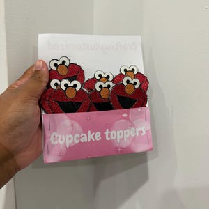 May include: A package of cupcake toppers featuring the character Elmo from Sesame Street. The toppers are red with black mouths, orange noses, and glitter accents. The package is pink and reads "Cupcake toppers".