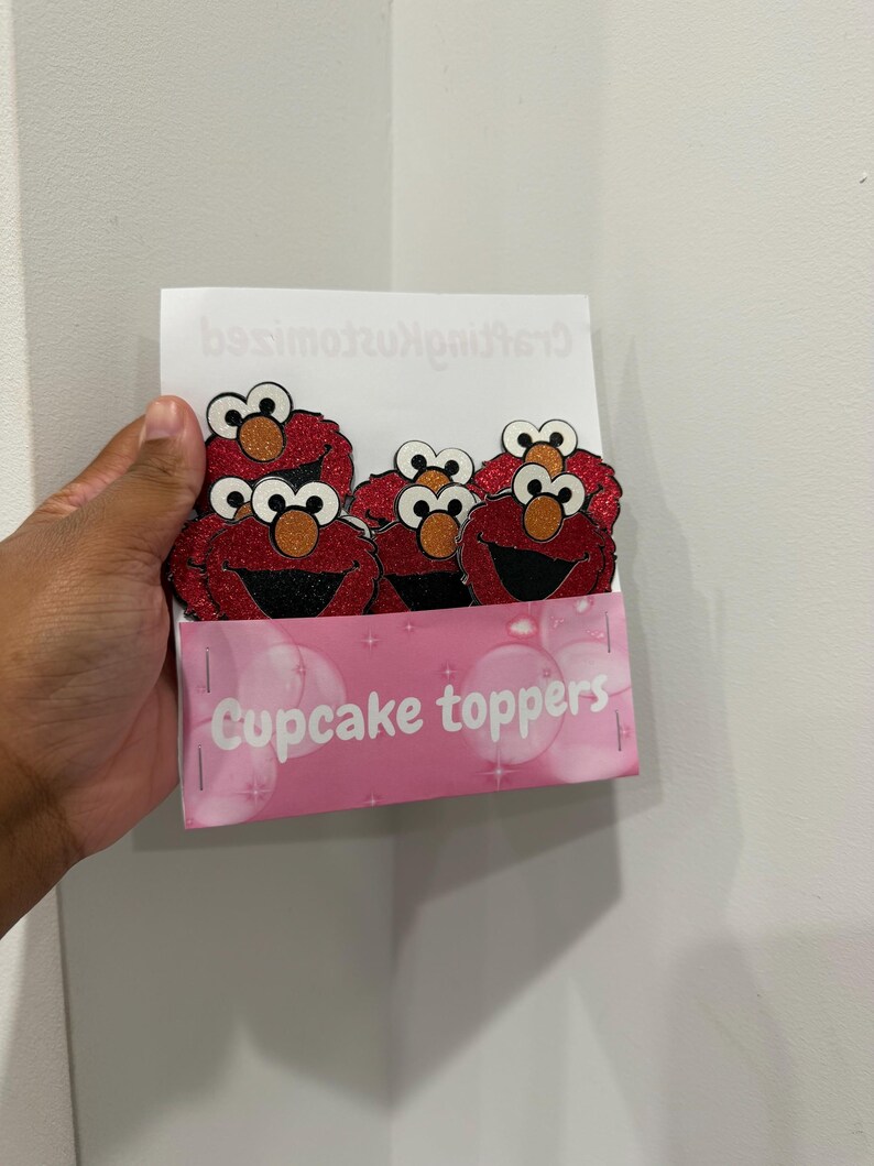 May include: A package of cupcake toppers featuring the character Elmo from Sesame Street. The toppers are red with black mouths, orange noses, and glitter accents. The package is pink and reads "Cupcake toppers".