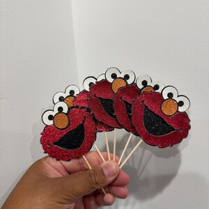May include: Red glitter Elmo face cupcake toppers with black mouths, orange noses, and white and black eyes. The toppers are attached to wooden sticks, perfect for decorating cupcakes or other baked goods.