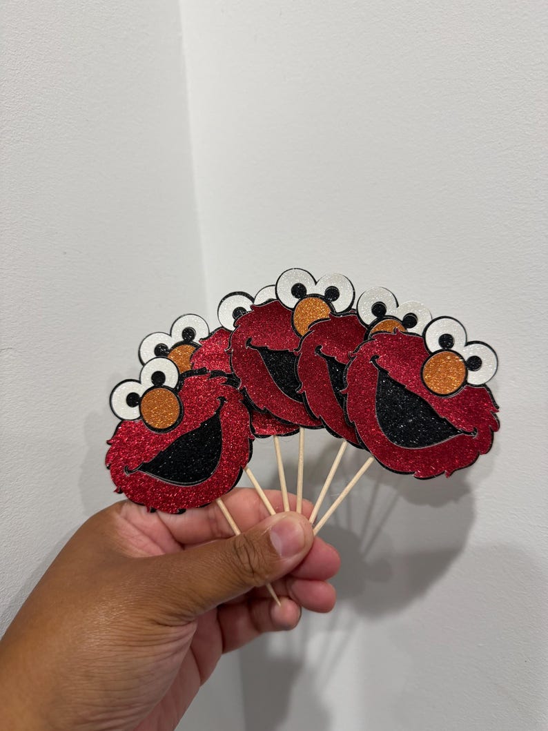 May include: Red glitter Elmo face cupcake toppers with black mouths, orange noses, and white and black eyes. The toppers are attached to wooden sticks, perfect for decorating cupcakes or other baked goods.