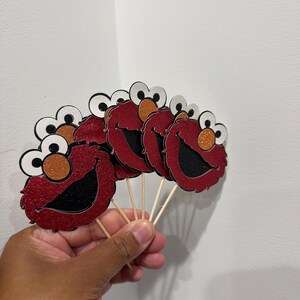 May include: Red glitter cupcake toppers featuring a cartoon character face with white eyes, orange nose, and a black mouth. The toppers are attached to wooden sticks, perfect for decorating cupcakes or other baked goods.