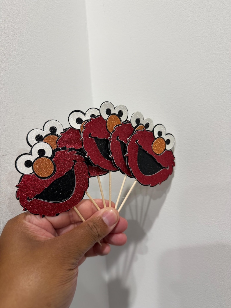 May include: Red glitter cupcake toppers featuring a cartoon character face with white eyes, orange nose, and a black mouth. The toppers are attached to wooden sticks, perfect for decorating cupcakes or other baked goods.