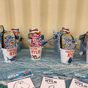 Puede incluir: Varios cubos de metal blancos con el texto "Finding Kylo" llenos de recuerdos de fiesta. Cada cubo presenta un personaje diferente de la película, incluyendo a Nemo y Dory. Los cubos están colocados sobre una mesa azul con una red de pesca.