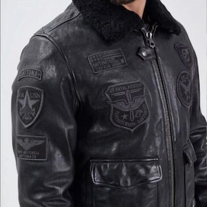 May include: Black leather bomber jacket with a black shearling collar. The jacket features multiple patches with text including "DAYTONA", "US NAVAL AVIATION", and "TOP GUN". It has two front pockets and a zipper closure.