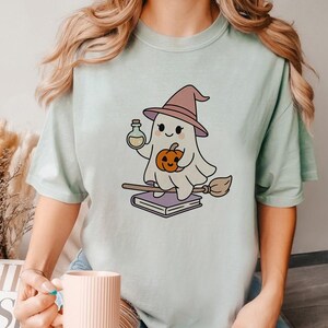 May include: A light green t-shirt featuring a cartoon ghost wearing a witch hat and holding a potion bottle and a pumpkin. The ghost is riding a broom and sitting on a stack of books. The design is in a cute, whimsical style.