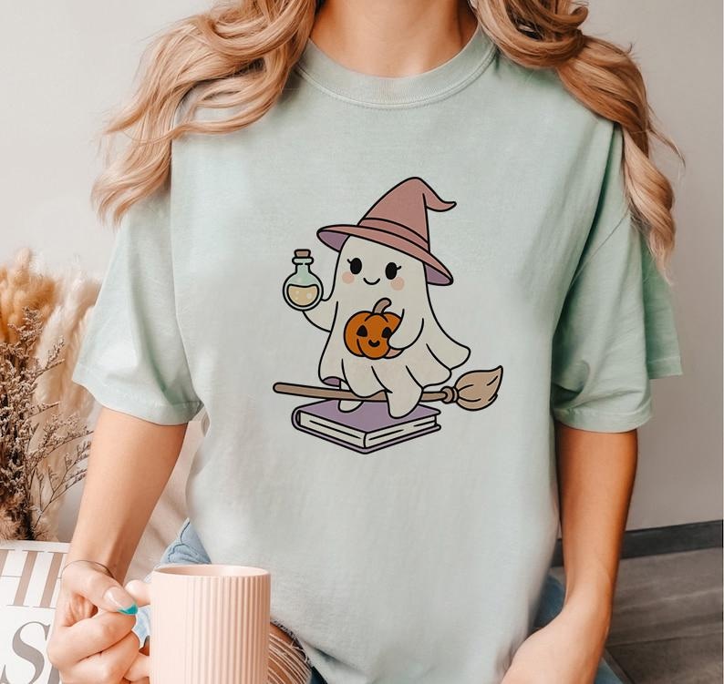 May include: A light green t-shirt featuring a cartoon ghost wearing a witch hat and holding a potion bottle and a pumpkin. The ghost is riding a broom and sitting on a stack of books. The design is in a cute, whimsical style.
