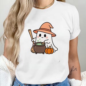 May include: White t-shirt featuring a cartoon ghost wearing an orange witch hat, holding a broom, and stirring a cauldron with green liquid. A small orange pumpkin sits beside the cauldron. Halloween-themed apparel.