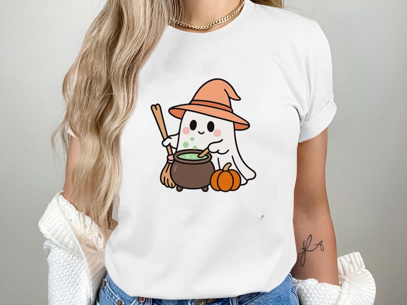 May include: White t-shirt featuring a cartoon ghost wearing an orange witch hat, holding a broom, and stirring a cauldron with green liquid. A small orange pumpkin sits beside the cauldron. Halloween-themed apparel.