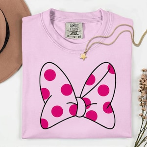 Pu&ograve; includere: Una maglietta rosa chiaro con un grande fiocco con pois magenta e un contorno nero. La maglietta ha uno scollo tondo e un'etichetta con la scritta "Comfort Colors". Una collana d'oro con un ciondolo a stella &egrave; drappeggiata sulla maglietta.