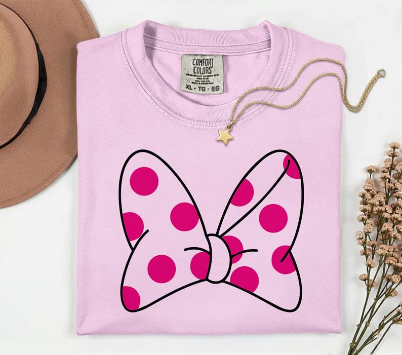 Pu&ograve; includere: Una maglietta rosa chiaro con un grande fiocco con pois magenta e un contorno nero. La maglietta ha uno scollo tondo e un'etichetta con la scritta "Comfort Colors". Una collana d'oro con un ciondolo a stella &egrave; drappeggiata sulla maglietta.