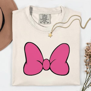 Pu&ograve; includere: T-shirt color crema con un grande fiocco rosa con contorno nero. La maglietta ha girocollo e maniche corte. Una collana d'oro con un ciondolo a forma di stella &egrave; appoggiata sulla maglietta. L'etichetta recita "COMFORT COLORS" e "XL - TG - EG".