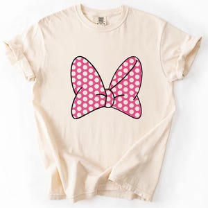Pu&ograve; includere: T-shirt color crema con un grande fiocco rosa a pois bianchi. Il fiocco &egrave; bordato di nero e centrato sul davanti della maglietta. La maglietta ha maniche corte e girocollo.