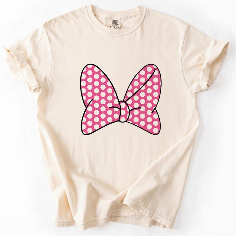 Pu&ograve; includere: T-shirt color crema con un grande fiocco rosa a pois bianchi. Il fiocco &egrave; bordato di nero e centrato sul davanti della maglietta. La maglietta ha maniche corte e girocollo.