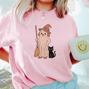 May include: Pink t-shirt featuring a cartoon dog dressed as a ghost, wearing a witch hat and holding a broom. A black cat sits beside the dog. The shirt is a light pink color, perfect for Halloween.