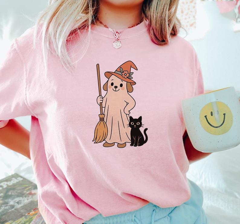 May include: Pink t-shirt featuring a cartoon dog dressed as a ghost, wearing a witch hat and holding a broom. A black cat sits beside the dog. The shirt is a light pink color, perfect for Halloween.