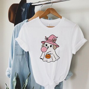 May include: White t-shirt featuring a cartoon dog dressed as a ghost for Halloween. The dog wears a pink witch hat and holds a pumpkin. The dog is blowing a pink bubble. The shirt is hanging on a wooden hanger.