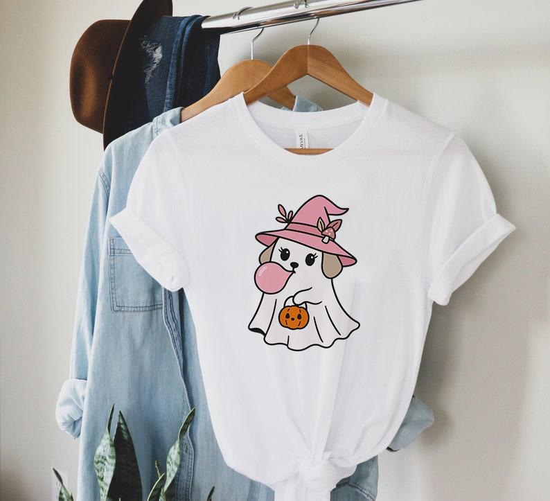 May include: White t-shirt featuring a cartoon dog dressed as a ghost for Halloween. The dog wears a pink witch hat and holds a pumpkin. The dog is blowing a pink bubble. The shirt is hanging on a wooden hanger.