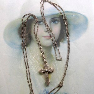May include: A gold-toned metal necklace featuring a delicate chain and an ornate cross pendant. The cross is decorated with intricate engraved patterns and a small, centered stone. The chain includes a small square link detail with a stone.