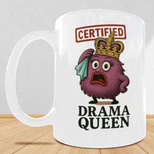 May include: White ceramic mug featuring a cartoon illustration of a purple creature wearing a gold crown and holding a cloth. The mug has the words "CERTIFIED" and "DRAMA QUEEN" printed on it.