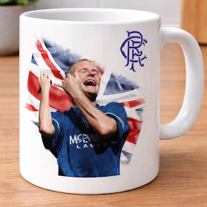 May include: A white ceramic mug featuring a graphic of a footballer in a blue jersey with the text McEwan Lager, positioned in front of a Union Jack flag. The blue Rangers Football Club crest is printed in the upper right corner of the mug.
