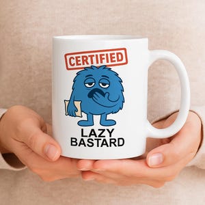 May include: A white ceramic mug featuring a graphic of a blue, furry, sleepy creature holding a small beige pillow. Above the creature is a red rectangular box with the text CERTIFIED, and below it is the black text LAZY BASTARD.