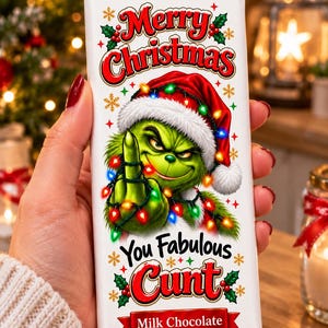 May include: A white milk chocolate bar wrapper featuring a green character wearing a Santa hat and wrapped in colourful Christmas lights. The text reads Merry Christmas You Fabulous Cunt in red and black lettering, decorated with holly and snowflakes.