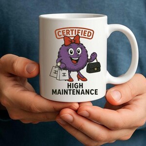May include: White ceramic mug with a cartoon character. The character has a purple body, red bow, and high heels. The mug has the text "CERTIFIED HIGH MAINTENANCE" and the character is holding shopping bags and a purse.