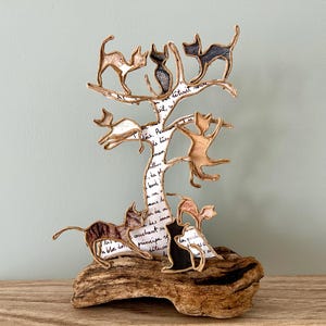 May include: A whimsical cat sculpture featuring several cat figures climbing a tree. The tree is made of paper with handwritten text, and the cats are in various poses. The sculpture is mounted on a piece of driftwood, creating a unique decorative piece.