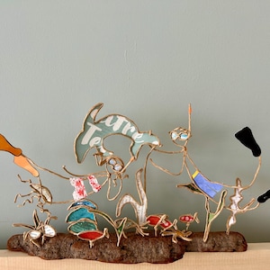 Puede incluir: Una escultura de alambre caprichosa que representa una escena de playa con un delfín, buceadores y varias criaturas marinas. La escultura incluye la palabra "Treasure" y está montada sobre un trozo de madera.