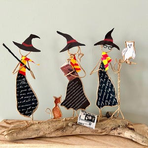 May include: Three whimsical wire witch figures with black hats, scarves, and dresses, standing on a piece of driftwood. One holds a wand, another a book, and the third an owl. A small orange cat and a newspaper are also present.