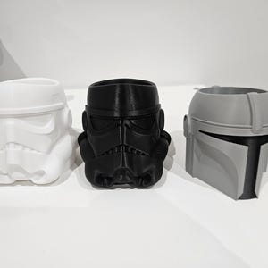 May include: Three character helmet planters, 3D-printed in white, black, and grey. The planters depict a Stormtrooper, an Imperial helmet, and a Mandalorian helmet. Ideal for small plants or decorative use.