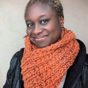 May include: An orange chunky knit infinity scarf. The scarf is made of a thick, textured yarn and is wrapped around the neck. The person in the image is wearing a black leather jacket and has short blonde hair.