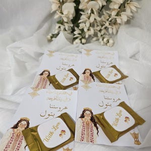 Puede incluir: Arreglo de tarjetas y bombones envueltos. Cada tarjeta presenta una ilustraci&oacute;n de una mujer con vestimenta tradicional, con texto &aacute;rabe. Los bombones est&aacute;n envueltos en papel dorado y tienen la inscripci&oacute;n "With Love".