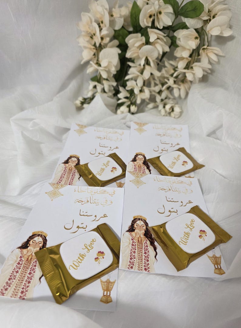Puede incluir: Arreglo de tarjetas y bombones envueltos. Cada tarjeta presenta una ilustraci&oacute;n de una mujer con vestimenta tradicional, con texto &aacute;rabe. Los bombones est&aacute;n envueltos en papel dorado y tienen la inscripci&oacute;n "With Love".