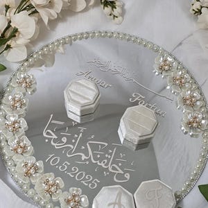 May include: A mirrored wedding ring tray with a pearl border and floral accents. The tray features two ring boxes and two initial boxes. The tray has the names "Anwar" and "Fartun" and the date "10.5.2025" engraved on it.