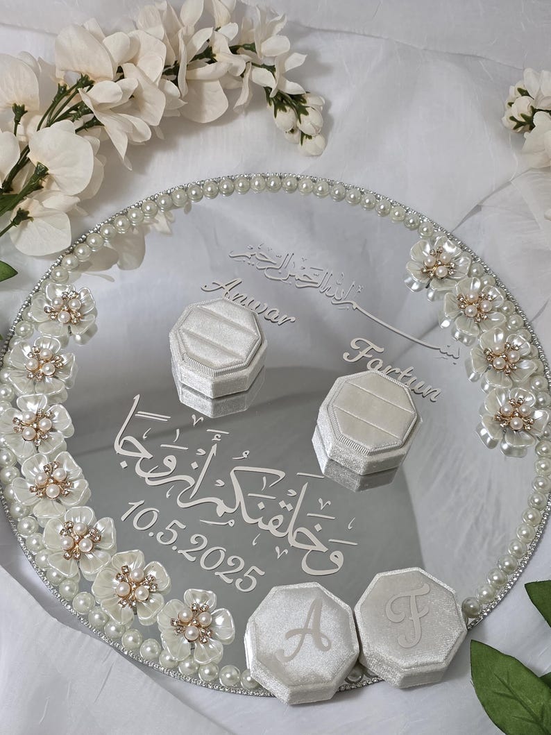 May include: A mirrored wedding ring tray with a pearl border and floral accents. The tray features two ring boxes and two initial boxes. The tray has the names "Anwar" and "Fartun" and the date "10.5.2025" engraved on it.
