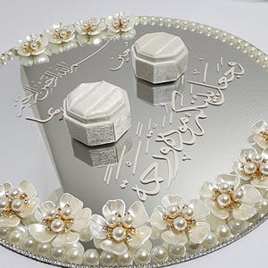 May include: A mirrored tray with a pearl border and floral accents. Two white ring boxes sit on the reflective surface, with decorative script. The tray is adorned with white flowers and pearls.