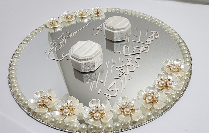 May include: A mirrored tray with a pearl border and floral accents. Two white ring boxes sit on the reflective surface, with decorative script. The tray is adorned with white flowers and pearls.