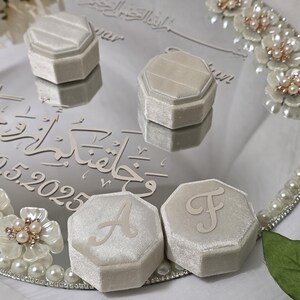May include: A mirrored tray with four light gray velvet ring boxes, two with the letters "A" and "F". The tray is decorated with pearls and floral accents. The tray has Arabic script and the date 05.2025.
