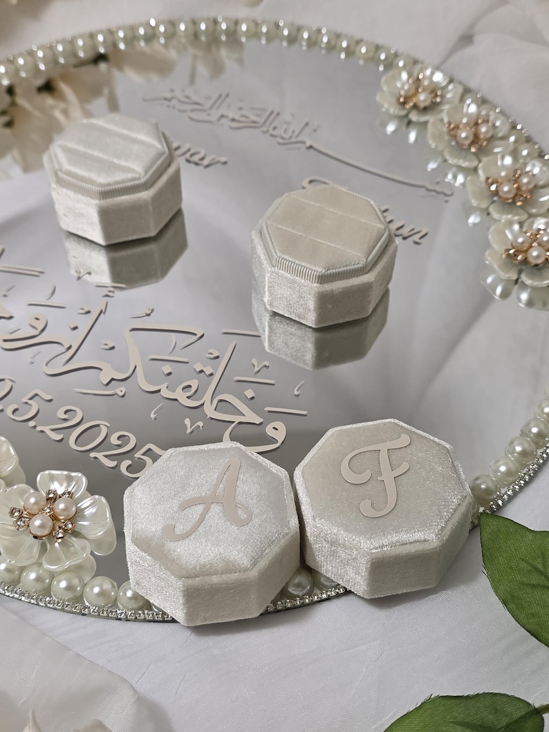 May include: A mirrored tray with four light gray velvet ring boxes, two with the letters "A" and "F". The tray is decorated with pearls and floral accents. The tray has Arabic script and the date 05.2025.