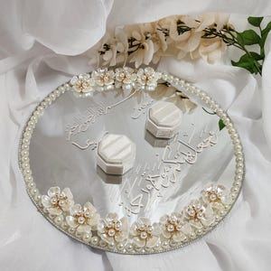 May include: A mirrored wedding ring tray with floral and pearl embellishments. The oval tray features white flowers with pearl centers and a pearl border. Two white ring boxes sit on the tray, which has Arabic script.