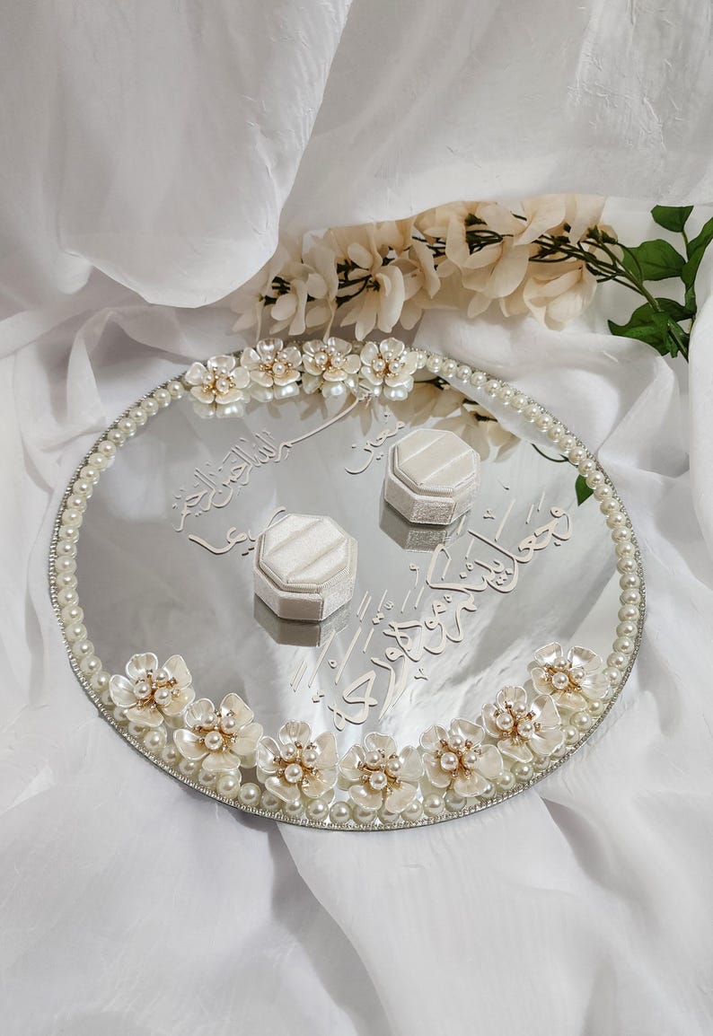 May include: A mirrored wedding ring tray with floral and pearl embellishments. The oval tray features white flowers with pearl centers and a pearl border. Two white ring boxes sit on the tray, which has Arabic script.